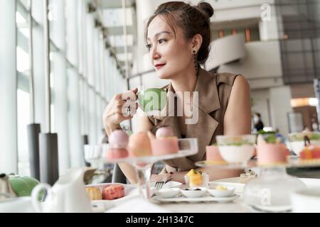 donna asiatica matura seduta al tavolo con tè pomeridiano nella caffetteria della lobby dell'hotel Foto Stock