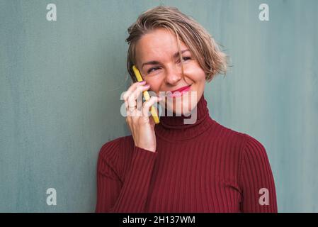 ritratto orizzontale di una donna bionda su un maglione di granata che parla per telefono mentre si appoggia su una parete verde Foto Stock