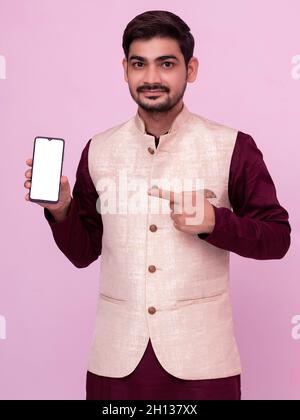 Ritratto di uomo con barba in costume tradizionale puntando al telefono cellulare e sorridendo alla macchina fotografica. Giovane uomo indiano in abbigliamento tradizionale e mostrando smartph Foto Stock