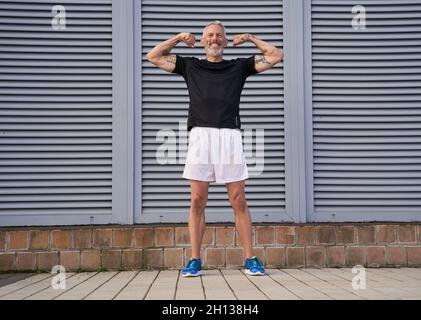 Scatto a tutta lunghezza di felice tatuato uomo di mezza età in buona forma sorridente mentre mostra le sue braccia muscolari, bicipite, posa all'aperto su sfondo grigio Foto Stock