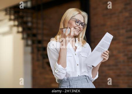 Approccio aziendale. Donna anziana elegante in occhiali e abbigliamento da lavoro sorridente, lettura di carte e parlare al telefono, in piedi a casa Foto Stock
