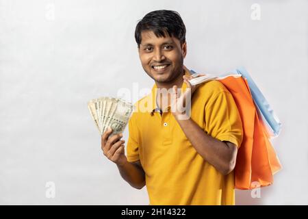 Un uomo indiano di mezza età in abito giallo con borse shopping e valute indiane sorridenti alla macchina fotografica su sfondo bianco Foto Stock