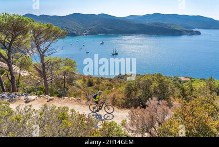 Bella donna che guida la sua mountain bike elettrica sulla costa del mediterraneo, sull'Isola d'Elba, nell'Arcipelago toscano, di fronte a Porto Foto Stock