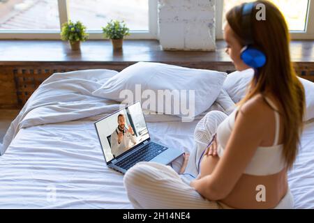 Foto di una donna incinta si siede a casa su un letto di fronte a un monitor portatile - consultazione online con un medico. Foto Stock