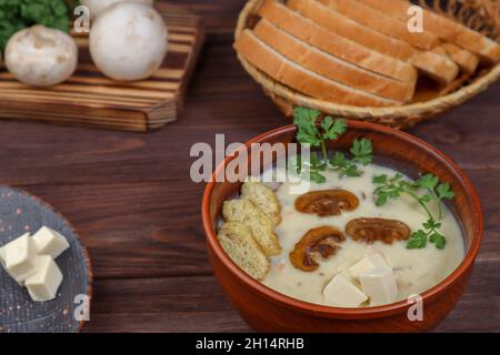 Zuppa di Champignon con formaggio fuso, prezzemolo e crostini in una ciotola primo piano su un tavolo di legno scuro sullo sfondo di un piatto con fuso Foto Stock