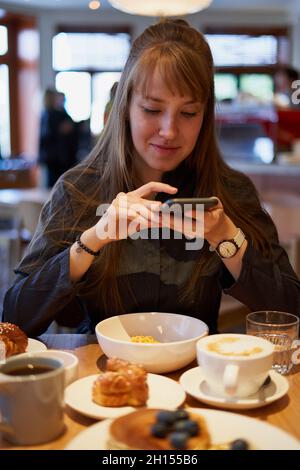 Donna che scatta foto di dolci e cappuccino con schiuma di latte al vapore in caffetteria Foto Stock