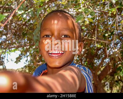 Capretto africano con una t-shirt blu nel cortile del villaggio in Botswana, su uno sfondo cespuglio, prendendo un selfie Foto Stock