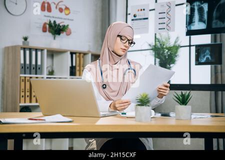 Fisioterapista femminile concentrato in occhiali e hijab leggere alcuni documenti mentre si siede in ufficio medico. Medico musulmano che utilizza un computer portatile wireless per il lavoro in ambienti chiusi. Foto Stock