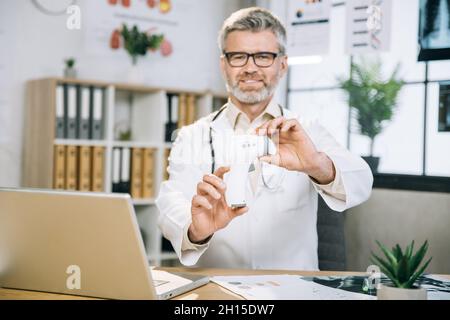 Medico maturo caucasico in camice da laboratorio bianco e occhiali seduti alla scrivania e mostrando sulla macchina fotografica moderno termometro elettronico. Specialista medico esperto che utilizza il computer portatile per lavorare in ufficio. Foto Stock