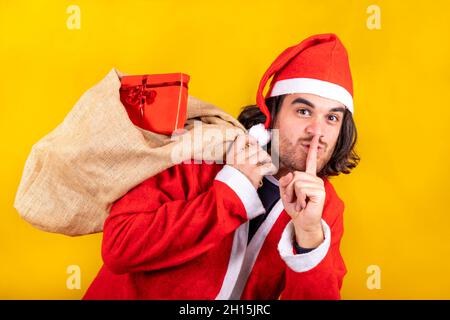 Un giovane bell'uomo vestito come Babbo Natale con un sacco pieno di regali invita a tacere e a tenere un segreto mettendo l'indice sulle sue labbra. Foto Stock