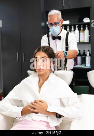 Parrucchiere con capelli grigi in maschera medica e occhiali lavaggio capelli di Donna in moderno salone Foto Stock