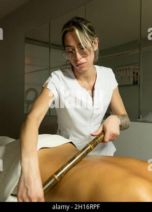 Massaggiatore femminile con bastone di bambù mentre massaggiano la parte posteriore del cliente in asciugamano sul divano in un salone di luce Foto Stock