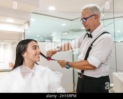 Cliente femminile sorridente in bianco mantello guardando maschile parrucchiere lavorando con i capelli Foto Stock