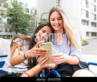 Donne dai capelli lunghi in gonne e bluse sorridenti e guardando lo schermo dello smartphone mentre si siede sulla panca in cortile Foto Stock