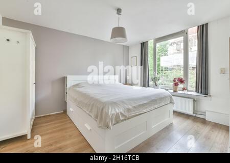 Interno della camera da letto arredato con letto coperto di bianco posto vicino armadio in legno sotto la lampada Foto Stock