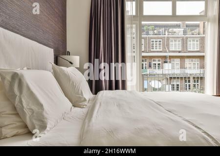 Letto confortevole e armadio in stile minimalista situato vicino alla finestra con tende nella camera da letto moderna Foto Stock