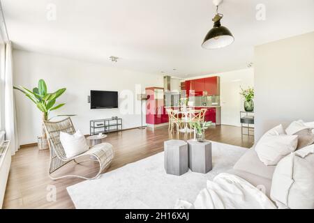 Interno di salotto con comodo divano e poltrona e cucina con mobili rossi in moderno appartamento open space Foto Stock