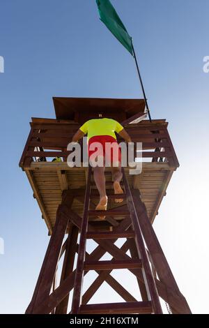 Vista posteriore ad angolo basso del corpo intero dell'irriconoscibile bagnino maschio che sale sulla scala sulla torre di salvataggio in legno sulla spiaggia Foto Stock