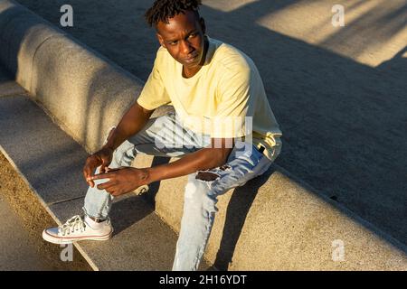 Corpo pieno di uomo nero con t-shirt casual e jeans e sneakers bianchi seduti sul bordo di cemento in strada alla luce del sole Foto Stock