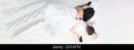 Uomo e donna sdraiati in camera da letto sul letto tenendo gli smartphone in mano e allontanandosi l'uno dall'altro Foto Stock