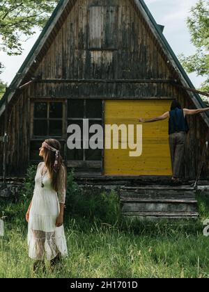 Primo piano di una ragazza attraente che guarda via e sullo sfondo un uomo che si bilancia sul portico di una casa di legno abbandonata Foto Stock