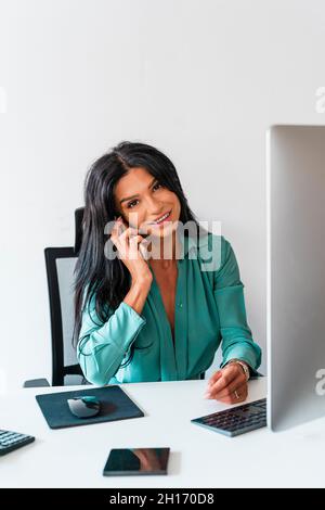 Sorridente dipendente femmina che ha una conversazione telefonica e guarda la fotocamera mentre si siede su sfondo bianco alla scrivania con il computer in un ufficio moderno Foto Stock