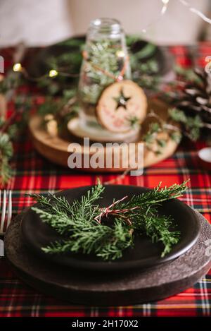 Tavolo di Natale con corona e ornamenti decorativi in legno e tovaglia a scacchi rossi con luci gialle sullo sfondo Foto Stock