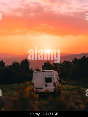 Caravan viaggiante bianco parcheggiato sul prato in altopiani contro il cielo arancione luminoso con il sole al tramonto in Galizia Foto Stock