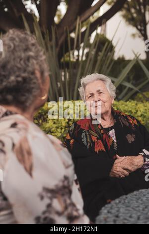 Vecchia femmina sorridente mentre si siede a tavola in un cortile esotico con palme e cespugli e godendo la conversazione con l'anziana amica femminile Foto Stock