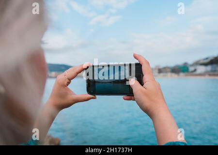 Vista laterale di donna elegante e irriconoscibile con capelli biondi che scattano foto dell'oceano blu contro il paesaggio urbano di Donostia su smartphone durante il giorno Foto Stock