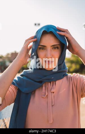 Positiva femmina musulmana che tocca il foulard tradizionale e guarda la macchina fotografica in giornata di sole in città Foto Stock