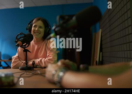 Happy femmina radio host in abiti casual e cuffie sorridenti durante la registrazione podcast in studio moderno con raccolto irriconoscibile collega Foto Stock
