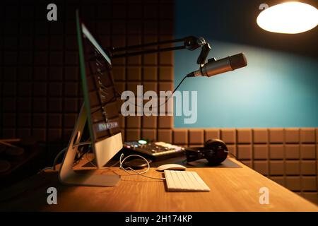 Computer e tastiera moderni posti su un tavolo di legno con microfono su treppiede e cuffie in studio di registrazione podcast scuro Foto Stock