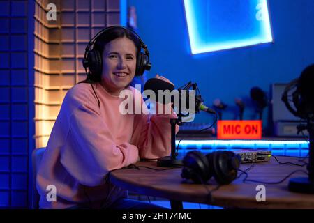 Allegro giovane ospite radio femmina sorridente e guardando la macchina fotografica mentre si siede in studio scuro con luci al neon al tavolo con microfoni e cuffie Foto Stock