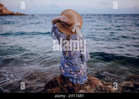 Vista posteriore di anonima giovane donna in abito estivo e cappello seduta su roccioso mare mentre si guarda via in estate sera Foto Stock