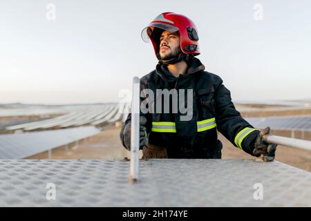Vigile del fuoco con casco protettivo rosso e uniforme in piedi sulla scala del camion dei vigili del fuoco e guardando lontano Foto Stock