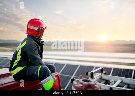 Anonimo Pensive adulto maschio indossando hardhat protettivo e tuta protettiva mentre si siede sulla parte superiore del motore di fuoco e guardando avanti ai pannelli solari in Foto Stock