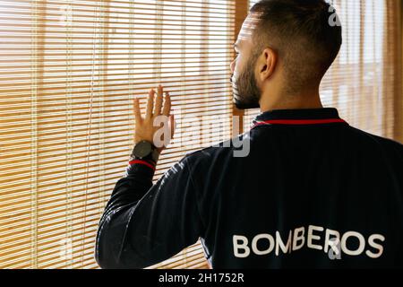 Vista laterale del raccolto di giovane etnia maschio portatore indossare giacca da sudore fireman guardando fuori finestra e toccando tende con le dita in luce solare brillante Foto Stock