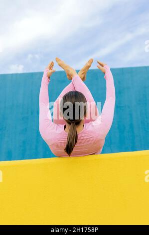Vista posteriore di una donna irriconoscibile in un abbigliamento sportivo rosa chiaro sdraiato sulla schiena con ginocchia al petto con gamba attraversata durante gli esercizi di yoga su blu e. Foto Stock