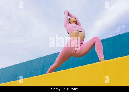 Da sotto di giovane donna che indossa abbigliamento sportivo in piedi in Virabhadrasana su parete gialla contro sfondo blu e cielo nuvoloso Foto Stock