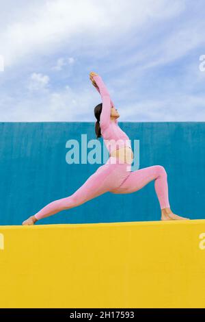 Da sotto di giovane donna che indossa abbigliamento sportivo in piedi in Virabhadrasana su parete gialla contro sfondo blu e cielo nuvoloso Foto Stock