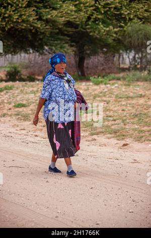 Vecchia donna bushman da Kalahari centrale, villaggio New Xade in Botswana, a piedi su una strada sterrata Foto Stock