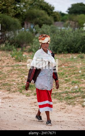 Vecchia donna bushman da Kalahari centrale, villaggio New Xade in Botswana, a piedi su una strada sterrata Foto Stock