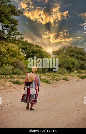 Vecchia donna bushman da Kalahari centrale, villaggio New Xade in Botswana, a piedi su una strada sterrata Foto Stock