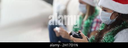 Donne in maschere mediche protettive e cappelli babbo natale seduti sul divano e tenendo console di gioco nelle loro mani a casa Foto Stock