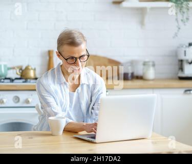 Felice donna di mezza età che scrive qualcosa sulla tastiera mentre lavora o studia sul laptop a casa Foto Stock