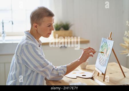 Donna creativa di mezza età che dipinge a casa mentre si siede a tavola di legno in cucina Foto Stock