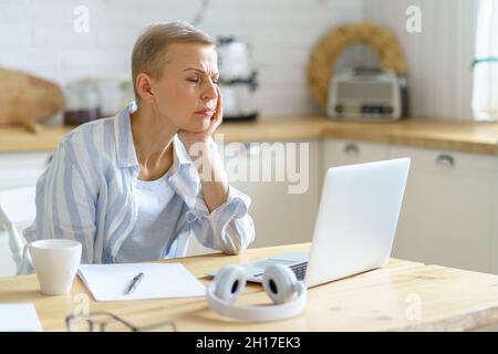 Donna anziana confusa che tiene la testa nelle mani e guarda sul laptop mentre lavora online da casa Foto Stock