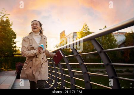 Bella donna allegra in trench beige gode di libertà, corre sul parco con un libro e takeaway carta tazza, sorrisi, sullo sfondo di belle bellezze Foto Stock