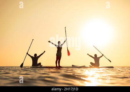 Happy Surfers si alza su tavole sup con braccia rialzate e guarda il tramonto Foto Stock